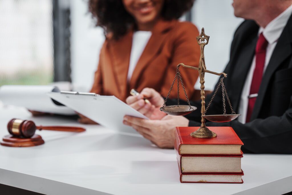 Scales of justice on a table with two lawyers discussing documents in the background.