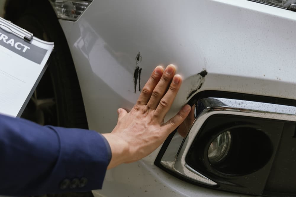 A person inspecting a scratch on a car's bumper while holding a clipboard with a contract.