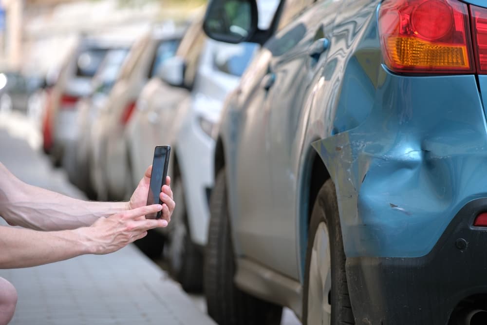 Person taking a photo of a blue car's dented rear side with a smartphone.