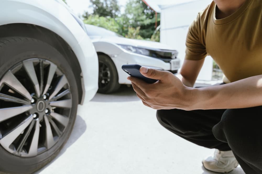 A person crouching next to a car tire, holding a smartphone, possibly documenting an accident.