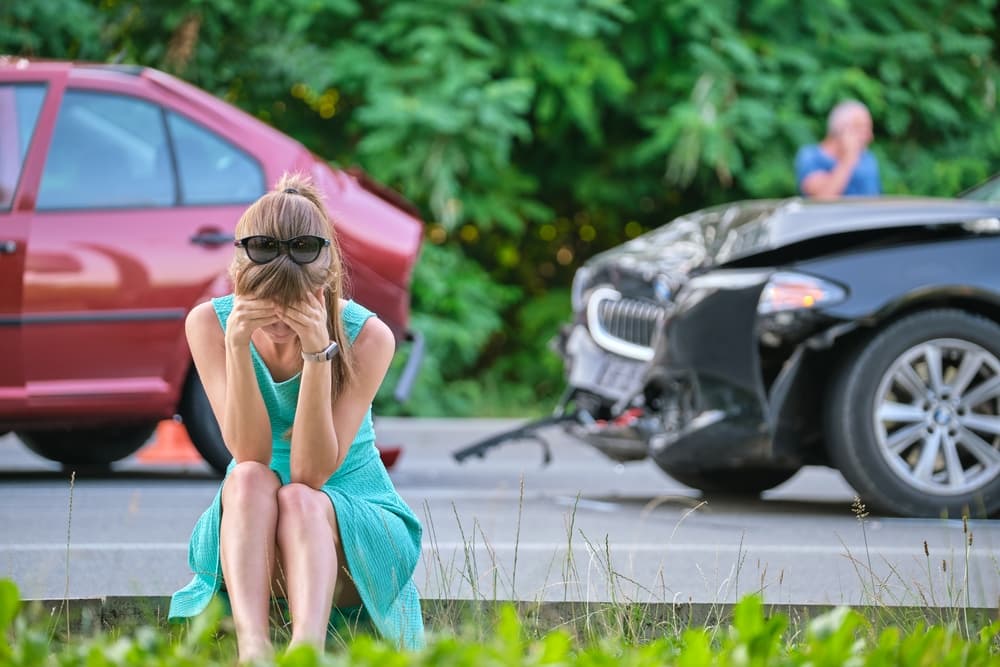 A distressed woman sitting on the roadside with two damaged cars in the background.