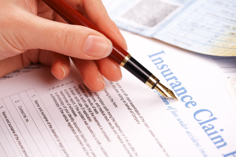 A hand completing an insurance claim form, with ID and vehicle documents visible in the background.