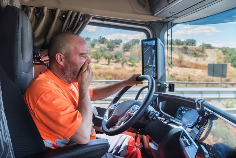 A truck driver is yawning while driving on the highway, clearly showing signs of tiredness and fatigue behind the wheel.