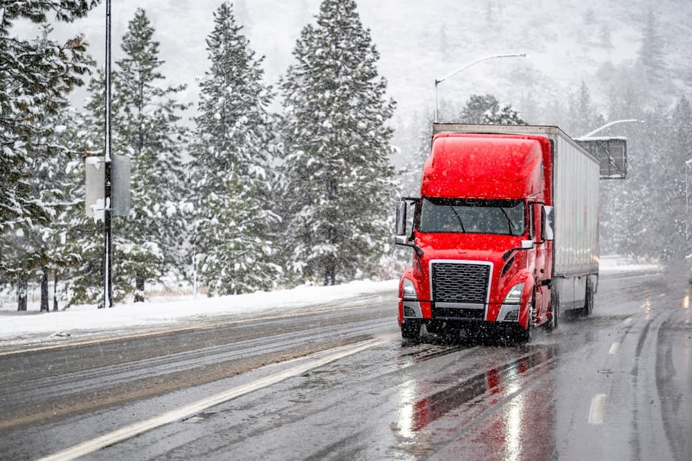 A large red big rig commercial semi-truck is transporting cargo in a dry van trailer, navigating a wet, winding road amidst a snow-covered winter forest during snowfall.






